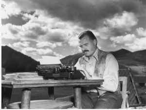 Black and white photo of classic authors Ernest Hemingway sitting at desk with mountains in the background