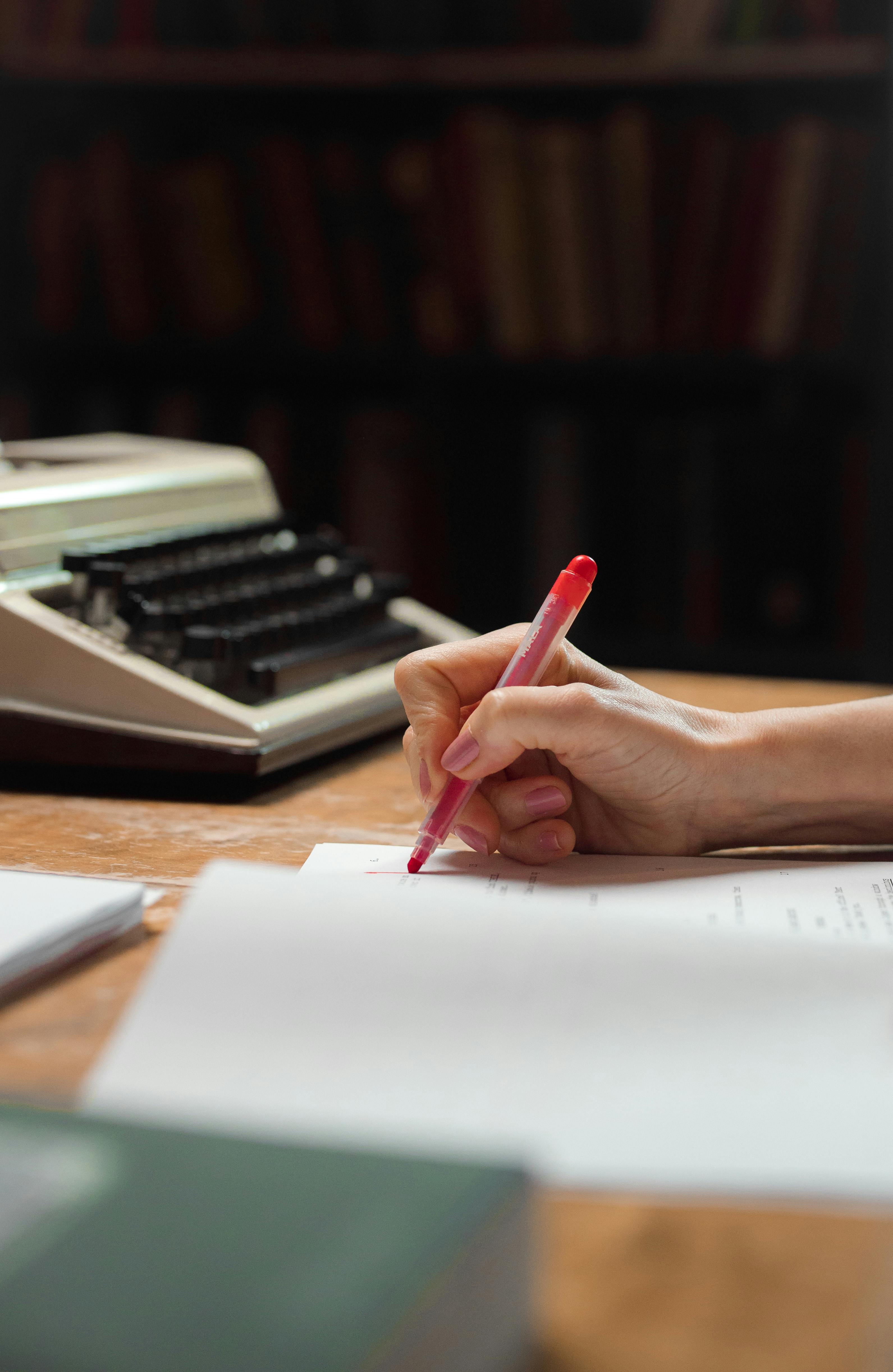Hand holding red pen for book proofreading with typewriter in the background.