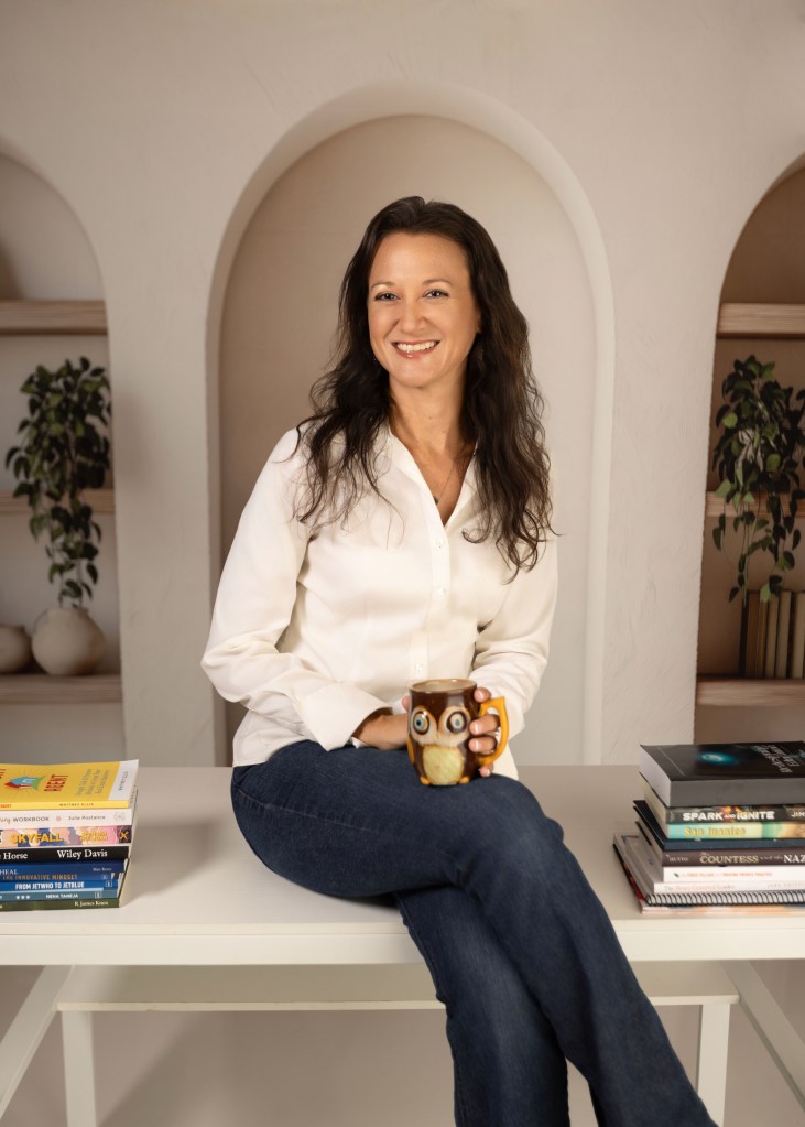Editor and writer Cortni Merritt, MA, sits on a desk with stacks of books she has edited around her.