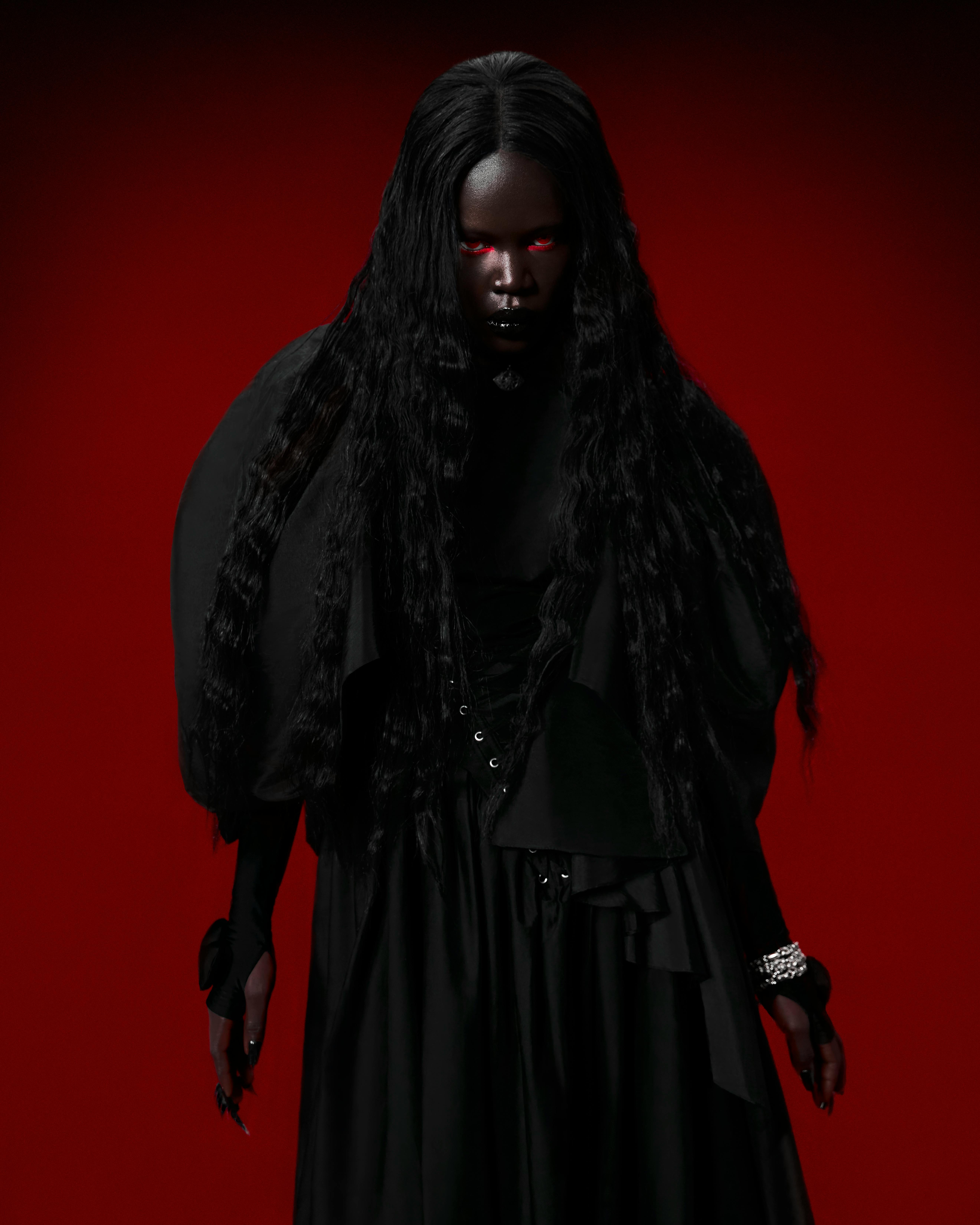 A dark-skinned woman with red eyes wearing all black on a red background stares menacingly into the camera.