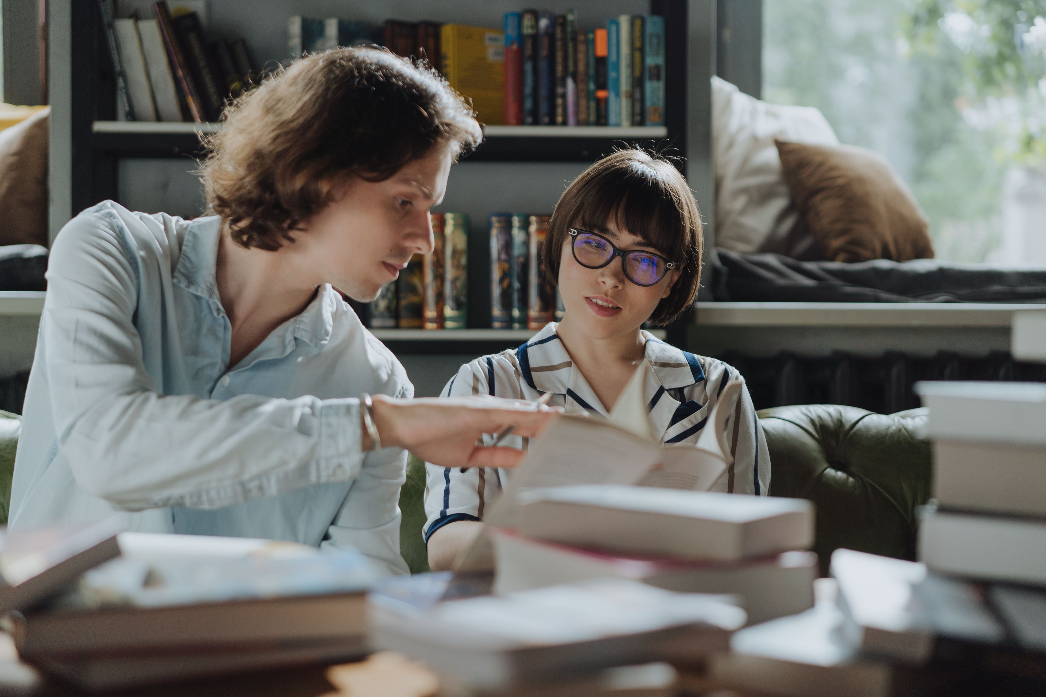 A writer and developmental book editor discussing a draft, shelves of books behind them and books scattered on the desk in front of them